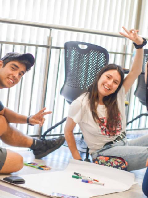 Un grupo de tres jóvenes sonrientes se sienta en el suelo, rodeados de materiales de dibujo en un ambiente interior luminoso.