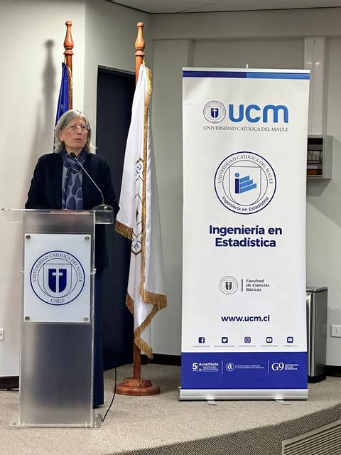 Una mujer está hablando en un podio frente a un banner de la Universidad Católica del Maule.