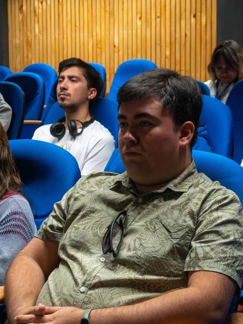 Un grupo de personas sentadas en un auditorio con sillas azules, prestando atención a una presentación.