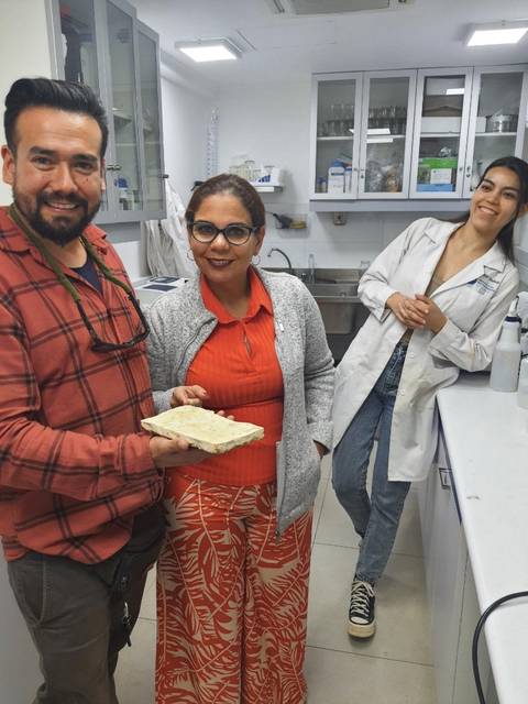 Tres personas sonrientes en un laboratorio, mostrando un recipiente con contenido en las manos.