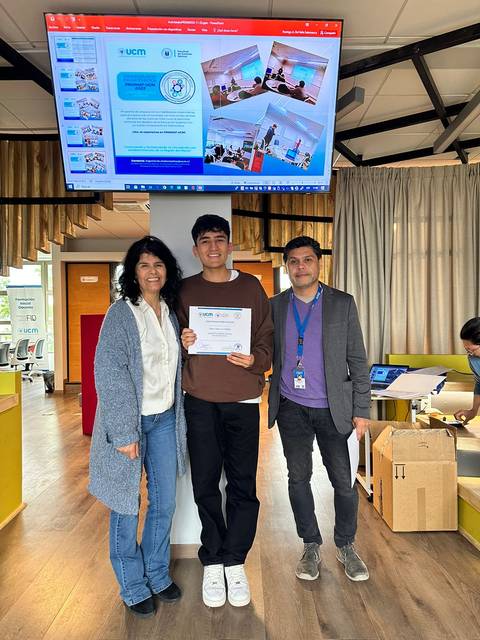 La imagen muestra a un grupo de tres personas en un ambiente académico, con una pantalla detrás que presenta información.