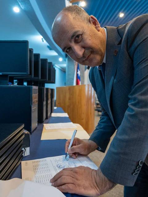 Un hombre firma documentos en una mesa frente a computadoras portátiles.