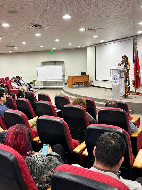 Una mujer habla desde un podio frente a una audiencia en un aula con asientos rojos.