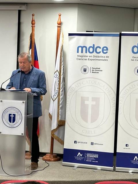 Un hombre está hablando en un podio frente a unas banderas y carteles de una universidad.