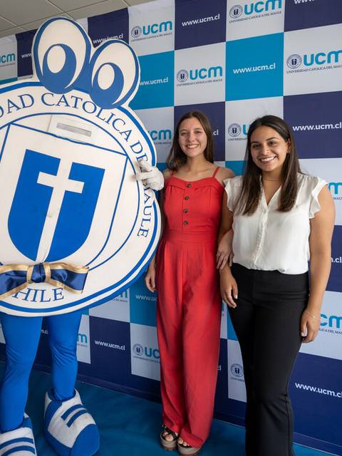 Dos mujeres posan junto a una mascota de la Universidad Católica del Maule en un evento.