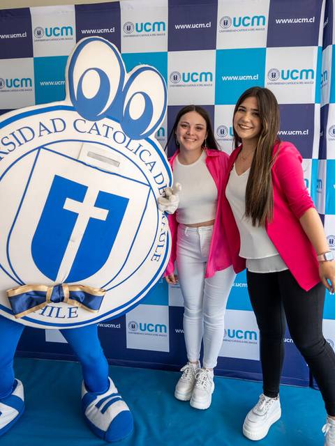 Dos mujeres posan junto a una mascota de la Universidad Católica del Maule en un evento.