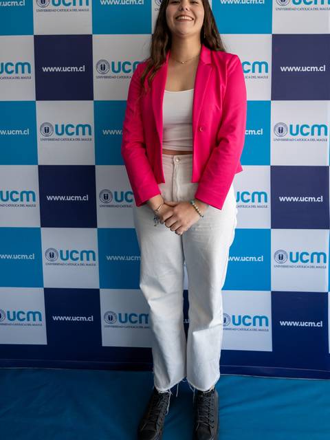 Una mujer sonriente viste una chaqueta rosa y pantalones blancos frente a un fondo con logotipos de la UCM.