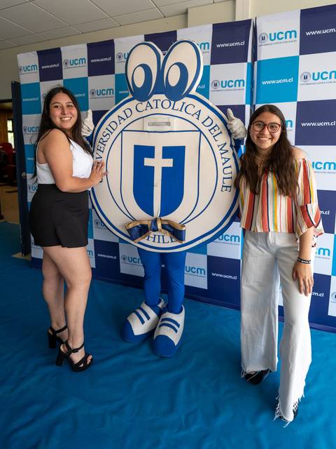 Dos mujeres posan junto a una mascota y un logo de la Universidad Católica del Maule en un evento.