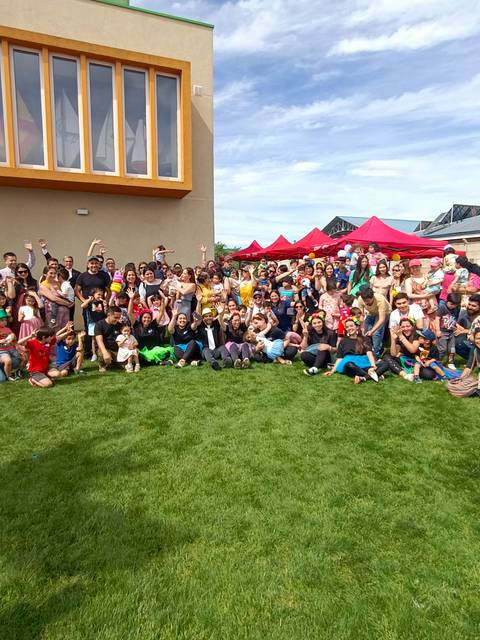 Un gran grupo de personas se reúne en un campo verde frente a un edificio, con carpas rojas al fondo.