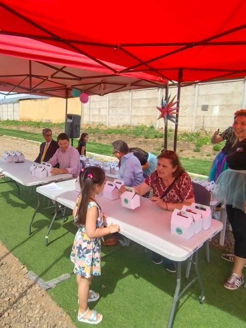 Un grupo de personas participa en un evento al aire libre bajo toldos, donde una niña se acerca a una mesa con suministros.
