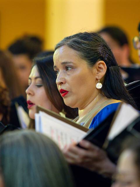 Un grupo de personas cantando en un coro, destacando a una mujer con pendientes y labios rojos que sostiene un libro de partituras.