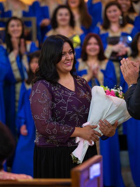 Una mujer sonriente sostiene un ramo de flores en un evento de celebración, mientras es aplaudida por un grupo de personas detrás de ella.