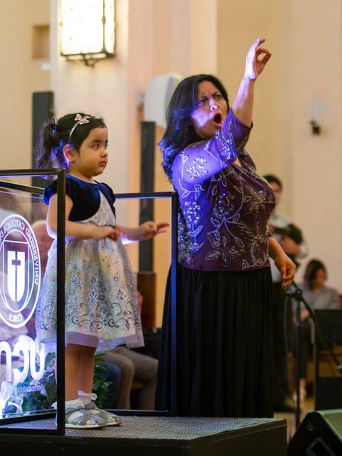 Una mujer y una niña están en el escenario de un evento religioso, la mujer está señalando hacia arriba mientras la niña la observa.
