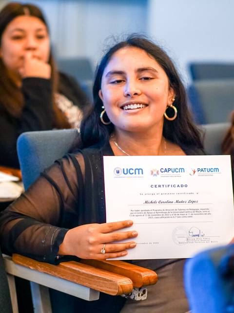Una joven sonriente sostiene un certificado en un evento con varios asistentes al fondo.