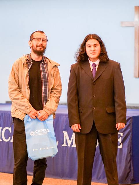 Dos hombres de pie en un escenario, uno con traje y el otro con chaqueta, frente a un fondo con una cruz.