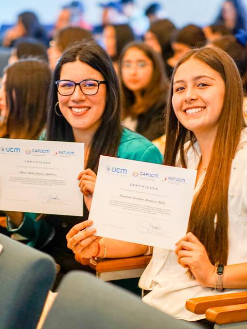 Dos jóvenes sonrientes sostienen certificados en un evento con un público de fondo.