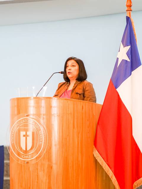 Una mujer habla desde un podio en una ceremonia oficial con banderas de Chile y la Universidad Católica de Chile en el fondo.