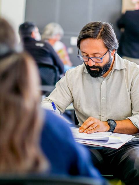 En la imagen se observa a un hombre con barba y gafas, concentrado en escribir en sus notas durante una reunión o taller.
