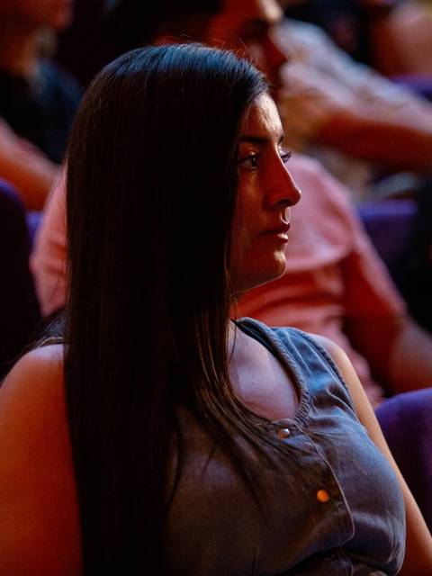 Una mujer con cabello largo y oscuro observa atentamente mientras está sentada en un auditorio.