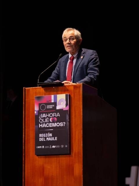 Un hombre de cabello canoso y gafas está hablando en un podio en un evento.
