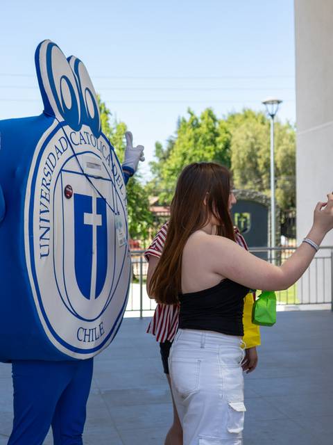 Un personaje disfrazado de una mano gigante interactúa con una persona que se está tomando una selfie en un evento universitario.