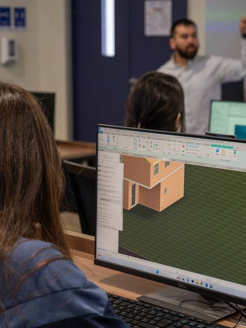 Un aula de informática donde se observa a un estudiante trabajando en un software de diseño arquitectónico mientras un instructor da una charla.