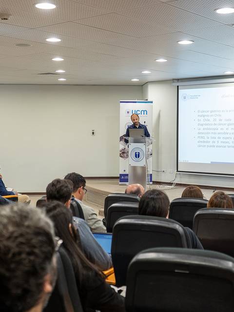 Un ponente presenta en una conferencia frente a un público atento en una sala con proyector y bandera chilena.
