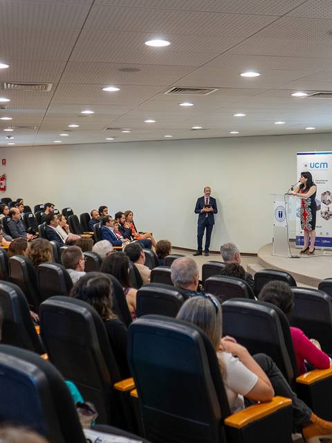 Una sala llena de gente asistiendo a una presentación con un orador y una presentadora frente a una pantalla al fondo.
