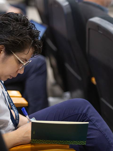 Una mujer toma notas en un cuaderno mientras escucha atentamente en un evento.