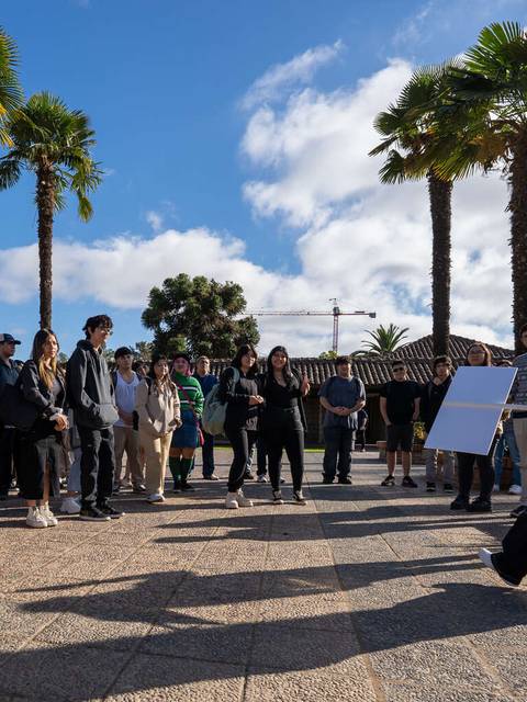 Un grupo de personas se reúne al aire libre, rodeado de palmeras, para escuchar a una presentadora que muestra un cartel.