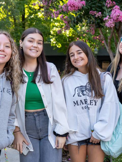 Cuatro amigas posando sonrientes en un entorno al aire libre rodeadas de flores.