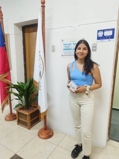 Una joven de pie sosteniendo un objeto frente a una pared decorada con banderas y una planta.