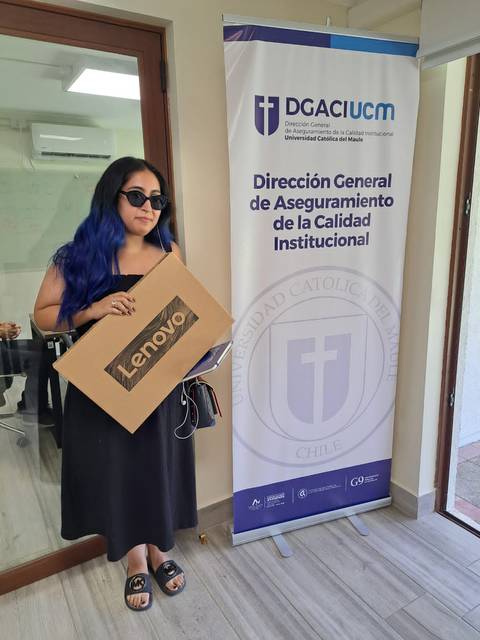Una persona con cabello azul sostiene una caja de Lenovo frente a un banner de la Universidad Católica de Chile.