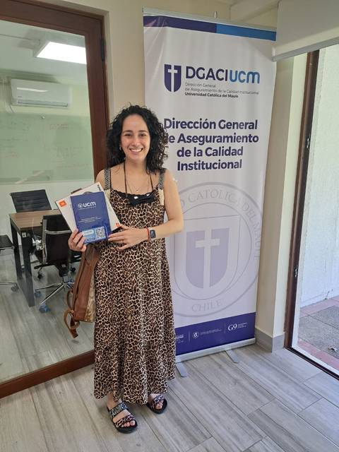 Una mujer sonriente sostiene documentos frente a una pared con un logo institucional.