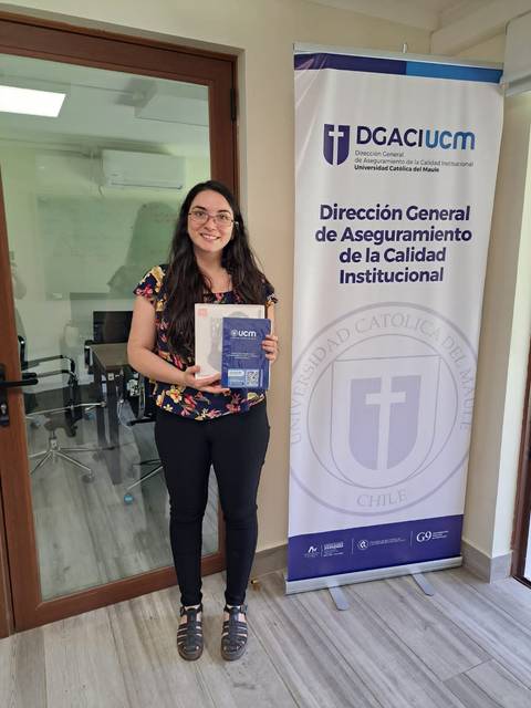 Una persona sonriente sostiene un documento frente a un panel informativo de la universidad.