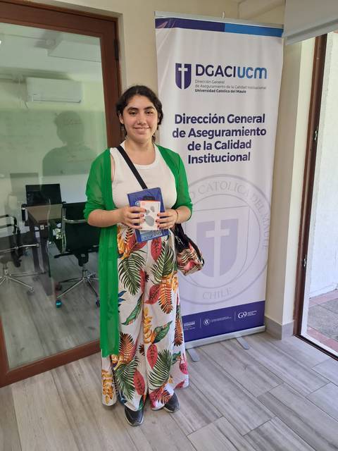 Una mujer de pie frente a un cartel de una institución educativa, sosteniendo un objeto en las manos.