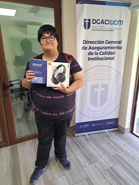 Un joven sonriente sostiene una caja de auriculares mientras está de pie en una oficina con un banner institucional de fondo.