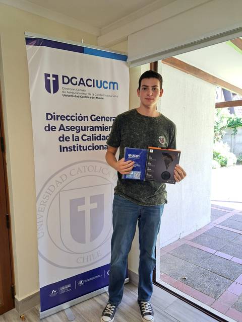 Un joven posa junto a un banner de una institución educativa mientras sostiene varios objetos en sus manos.
