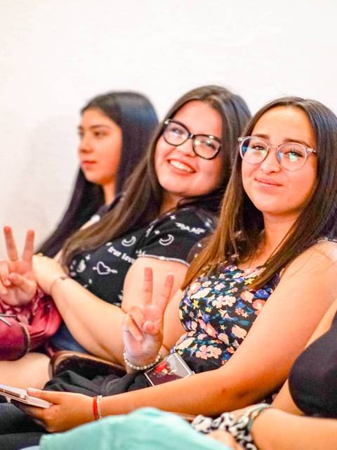 Un grupo de cuatro jóvenes sonrientes sentadas, mostrando gestos de paz y vestidas con ropa colorida.