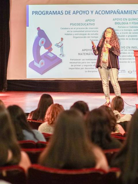 Una presentadora habla sobre programas de apoyo educativo frente a una audiencia en un auditorio.