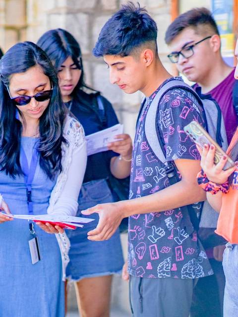 Un grupo de jóvenes se encuentra en un lugar al aire libre, interactuando mientras revisan documentos.