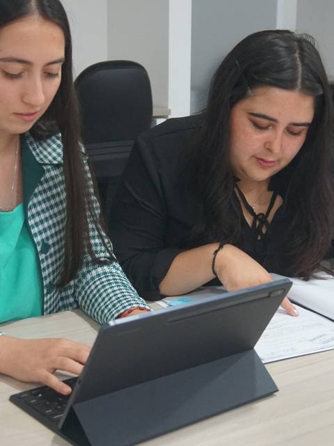 Dos mujeres están trabajando juntas en una mesa, una utilizando una computadora portátil y la otra revisando documentos.