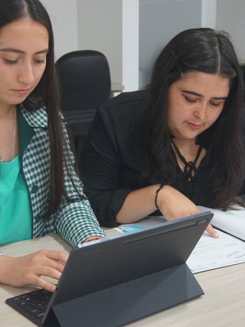Dos mujeres estudiantes trabajan juntas en un ambiente de oficina con documentos y una computadora portátil.