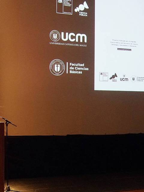 Un presentador habla en un escenario con una mesa y una pantalla que muestra un cartel de un evento sobre 'autopoyética'.