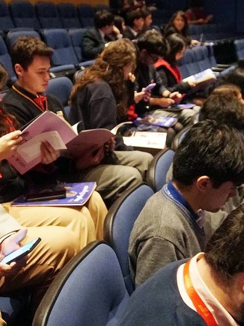 Un grupo de personas sentadas en un auditorio usando dispositivos móviles y revisando materiales impresos.