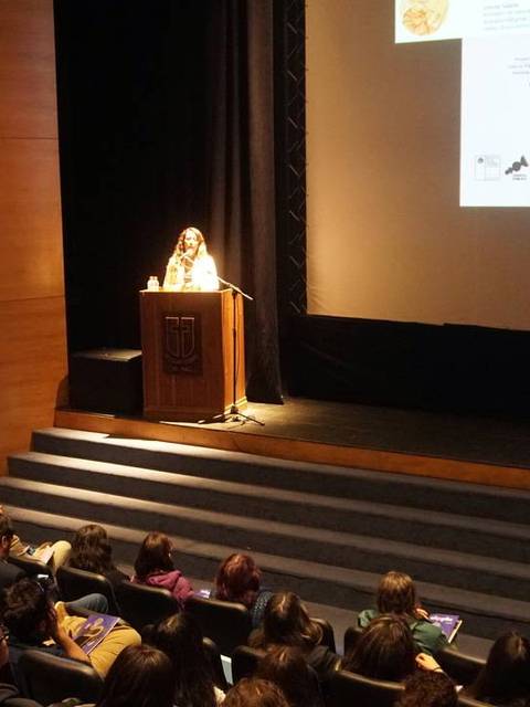 Una oradora presenta en un auditorio mientras el público escucha atentamente.