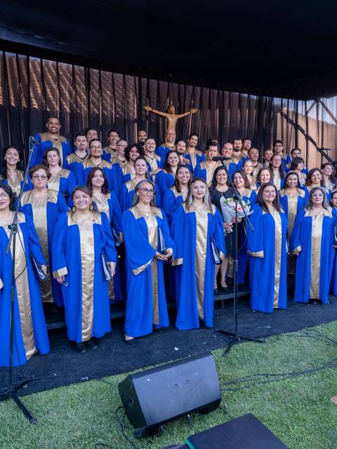 Un coro compuesto por numerosos cantantes, todos vestidos con túnicas azules y doradas, posando en un escenario al aire libre.