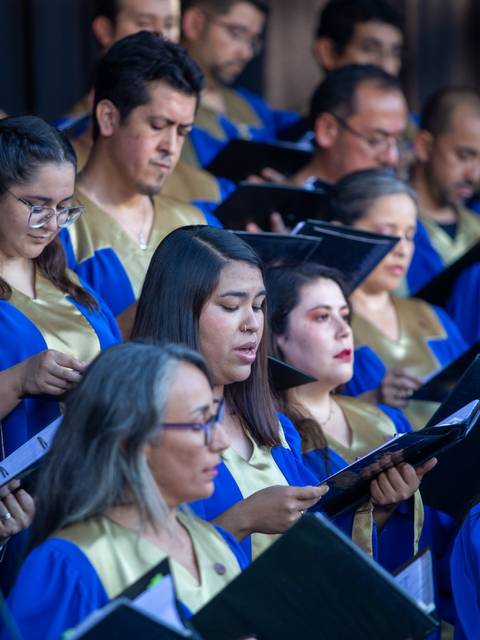 Un coro de personas en un escenario, vistiendo túnicas azules y doradas, mientras cantan y sostienen partituras.