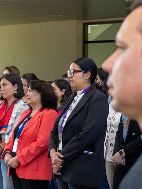 Un grupo de personas de pie en un evento, escuchando atentamente.
