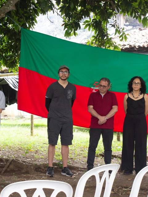 Un grupo de personas participan en un evento al aire libre bajo un árbol, con banderas de fondo.